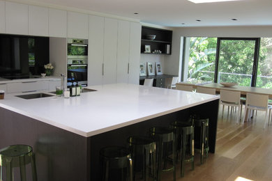Design ideas for a large modern single-wall eat-in kitchen in Melbourne with an undermount sink, white cabinets, solid surface benchtops, green splashback, glass sheet splashback, stainless steel appliances, light hardwood floors and with island.