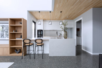 This is an example of a modern kitchen in Canberra - Queanbeyan with a built-in sink, recessed-panel cabinets, white cabinets, engineered stone countertops, white splashback, engineered quartz splashback, black appliances, concrete flooring, an island, grey floors, white worktops and a timber clad ceiling.