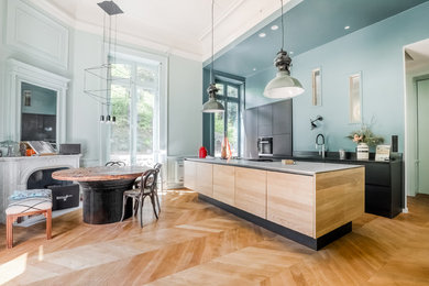 This is an example of a large modern single-wall eat-in kitchen in Lyon with an undermount sink, beaded inset cabinets, light wood cabinets, black splashback, marble splashback, black appliances, light hardwood floors, with island, beige floor and black benchtop.