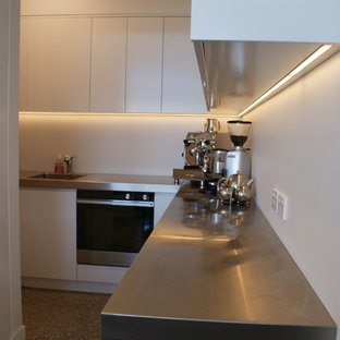 Large industrial kitchen pantry pictures - Inspiration for a large industrial galley concrete floor kitchen pantry remodel in Dunedin with an integrated sink, flat-panel cabinets, white cabinets, stainless steel countertops, white backsplash, glass sheet backsplash, stainless steel appliances and an island