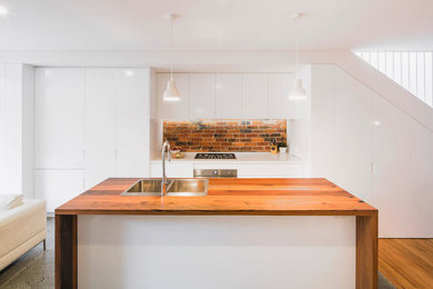 Photo of a mid-sized contemporary galley kitchen pantry in Sydney with a double-bowl sink, white cabinets, wood benchtops, brown splashback, brick splashback, stainless steel appliances, medium hardwood floors, with island and brown floor.
