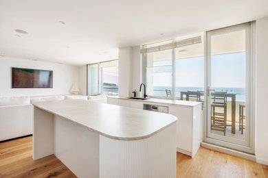 Photo of a mid-sized beach style u-shaped open plan kitchen in Sydney with an undermount sink, flat-panel cabinets, white cabinets, quartzite benchtops, porcelain splashback, black appliances, medium hardwood floors and with island.