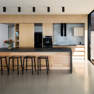 Large contemporary galley kitchen in Sunshine Coast with an undermount sink, flat-panel cabinets, light wood cabinets, grey splashback, black appliances, with island, grey floor and black benchtop.