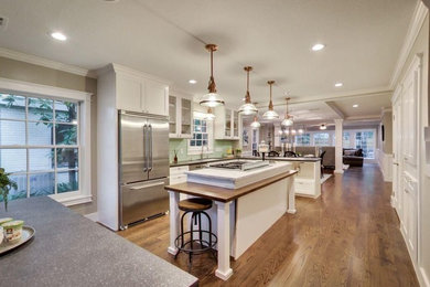 Eat-in kitchen - mid-sized transitional l-shaped medium tone wood floor and brown floor eat-in kitchen idea in Other with an undermount sink, glass-front cabinets, white cabinets, solid surface countertops, green backsplash, subway tile backsplash, stainless steel appliances and an island