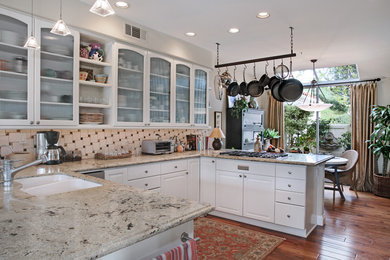 Design ideas for a mid-sized eclectic u-shaped open plan kitchen in Orange County with an undermount sink, glass-front cabinets, white cabinets, granite benchtops, beige splashback, glass tile splashback, stainless steel appliances, medium hardwood floors and no island.