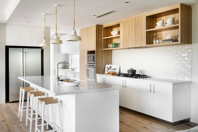 Photo of a beach style l-shaped kitchen in Perth with white splashback, ceramic splashback, an undermount sink, flat-panel cabinets, white cabinets, stainless steel appliances, medium hardwood floors and with island.