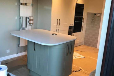 This is an example of a large contemporary l-shaped open plan kitchen in Other with a single-bowl sink, shaker cabinets, green cabinets, solid surface benchtops, grey splashback, ceramic splashback, black appliances, vinyl floors, with island, brown floor and white benchtop.