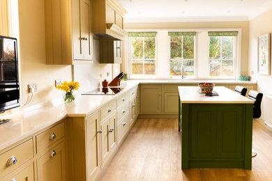 Inspiration for a medium sized country l-shaped enclosed kitchen in Essex with a double-bowl sink, shaker cabinets, beige cabinets, engineered stone countertops, white splashback, engineered quartz splashback, black appliances, light hardwood flooring, an island, brown floors, white worktops and a chimney breast.
