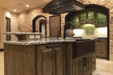 Photo of a mid-sized mediterranean eat-in kitchen in Austin with a farmhouse sink, raised-panel cabinets, distressed cabinets, granite benchtops, beige splashback, stone tile splashback, stainless steel appliances, travertine floors and with island.