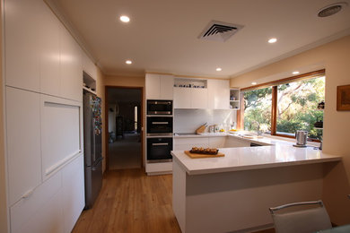 Mid-sized contemporary u-shaped open plan kitchen in Melbourne with a single-bowl sink, glass-front cabinets, white cabinets, granite benchtops, white splashback, stone tile splashback, black appliances, linoleum floors, a peninsula, beige floor and white benchtop.