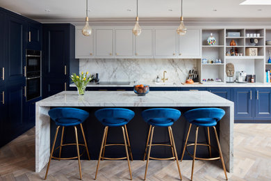 Design ideas for a large transitional l-shaped kitchen in London with blue cabinets, marble benchtops, white splashback, marble splashback, panelled appliances, light hardwood floors, with island, white benchtop, an undermount sink, recessed-panel cabinets and beige floor.
