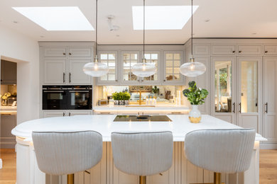 Design ideas for a medium sized classic galley kitchen/diner in London with an integrated sink, shaker cabinets, beige cabinets, quartz worktops, metallic splashback, mirror splashback, light hardwood flooring, an island and white worktops.