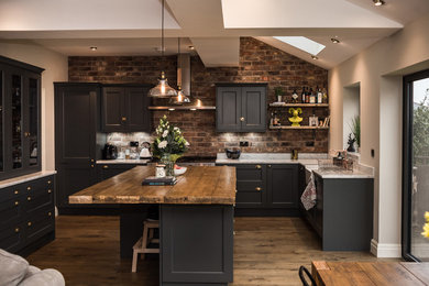Inspiration for a mid-sized traditional l-shaped open plan kitchen in Cheshire with a farmhouse sink, with island, shaker cabinets, black cabinets, brick splashback, medium hardwood floors and marble benchtops.