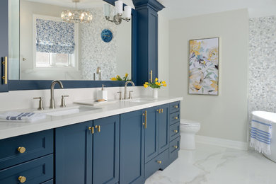Design ideas for a large transitional master bathroom in Toronto with blue cabinets, a freestanding tub, white tile, marble, grey walls, ceramic floors, engineered quartz benchtops, white floor, white benchtops, recessed-panel cabinets and an undermount sink.