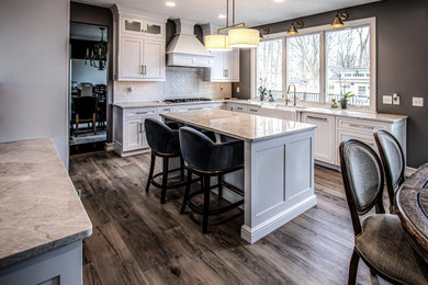 Example of a mid-sized transitional u-shaped vinyl floor and brown floor eat-in kitchen design in Cleveland with a farmhouse sink, flat-panel cabinets, white cabinets, quartz countertops, white backsplash, ceramic backsplash, stainless steel appliances, an island and beige countertops
