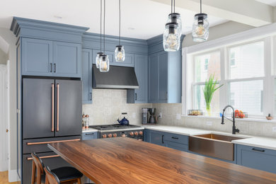Example of a mid-sized cottage l-shaped medium tone wood floor and brown floor eat-in kitchen design in Boston with a farmhouse sink, recessed-panel cabinets, blue cabinets, wood countertops, beige backsplash, ceramic backsplash, black appliances, an island and brown countertops