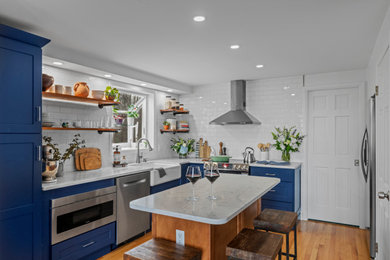 Design ideas for a transitional l-shaped kitchen in Providence with a farmhouse sink, shaker cabinets, blue cabinets, white splashback, ceramic splashback, stainless steel appliances, medium hardwood floors, with island and white benchtop.