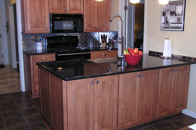 Inspiration for a small timeless galley porcelain tile and brown floor eat-in kitchen remodel in Kansas City with a farmhouse sink, shaker cabinets, medium tone wood cabinets, granite countertops, metallic backsplash, stone tile backsplash, black appliances and a peninsula