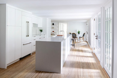 На фото: прямая кухня-гостиная среднего размера в современном стиле с плоскими фасадами, белыми фасадами, белым фартуком, паркетным полом среднего тона, островом, бежевым полом и белой столешницей