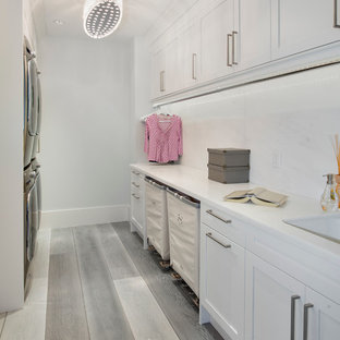 Photo of a classic galley utility room in Miami with shaker cabinets, white cabinets, a stacked washer and dryer, white walls and light hardwood flooring.