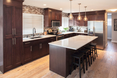 Large contemporary l-shaped kitchen in Calgary with shaker cabinets, stainless steel appliances, an undermount sink, dark wood cabinets, quartz benchtops, brown splashback, stone tile splashback, light hardwood floors, with island and brown floor.