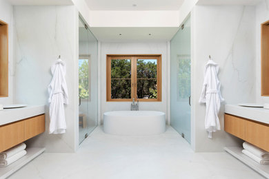 Свежая идея для дизайна: большая главная ванная комната в стиле модернизм с светлыми деревянными фасадами, отдельно стоящей ванной, душем без бортиков, плиткой из листового камня, белыми стенами, мраморным полом, мраморной столешницей, белым полом, душем с распашными дверями, белой столешницей, подвесной тумбой, плоскими фасадами, белой плиткой и тумбой под две раковины - отличное фото интерьера