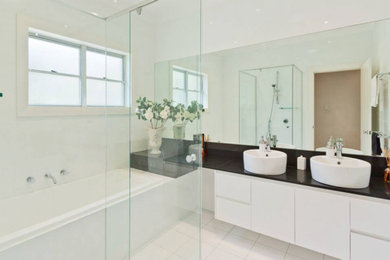 Mid-sized contemporary master bathroom in Sydney with flat-panel cabinets, white cabinets, a drop-in tub, an alcove shower, a two-piece toilet, white tile, ceramic tile, white walls, porcelain floors, a vessel sink, engineered quartz benchtops, white floor, a hinged shower door and black benchtops.
