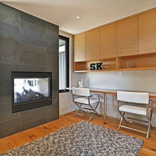 This is an example of a large modern home office and library in Toronto with white walls, light hardwood flooring, a standard fireplace, a tiled fireplace surround, a built-in desk and brown floors.