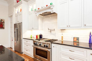 Example of a large transitional u-shaped medium tone wood floor and multicolored floor open concept kitchen design in Other with a farmhouse sink, recessed-panel cabinets, white cabinets, quartz countertops, white backsplash, subway tile backsplash, stainless steel appliances, an island and black countertops