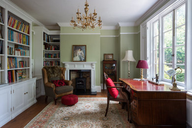 Klassisches Arbeitszimmer mit Arbeitsplatz, grüner Wandfarbe, dunklem Holzboden, Kaminofen, Kaminumrandung aus Holz, freistehendem Schreibtisch und braunem Boden in Buckinghamshire