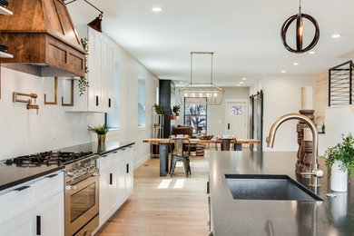 Eat-in kitchen - mid-sized cottage galley light wood floor and beige floor eat-in kitchen idea in Denver with an undermount sink, shaker cabinets, white cabinets, white backsplash, ceramic backsplash, stainless steel appliances, an island and black countertops