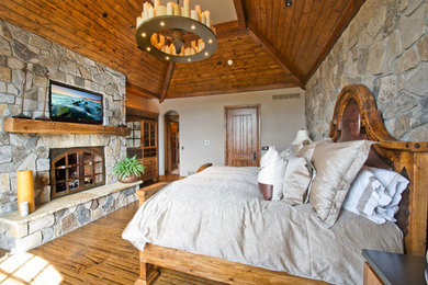Example of a large mountain style master medium tone wood floor and brown floor bedroom design in Denver with gray walls, a standard fireplace and a stone fireplace