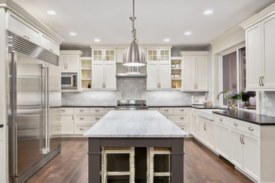 Photo of a large country u-shaped separate kitchen in Atlanta with shaker cabinets, white cabinets, quartz benchtops, white splashback, with island, white benchtop, a farmhouse sink, porcelain splashback, stainless steel appliances, medium hardwood floors and brown floor.