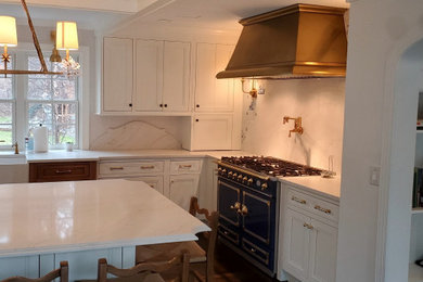 Example of a huge transitional l-shaped coffered ceiling eat-in kitchen design in New York with a farmhouse sink, recessed-panel cabinets, white cabinets, white backsplash, stone slab backsplash, colored appliances, an island and white countertops