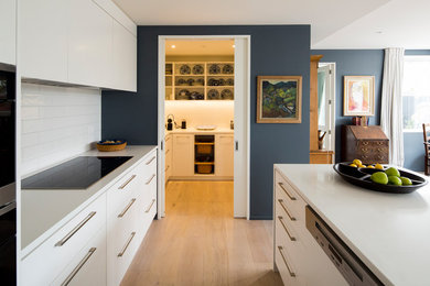 Mid-sized contemporary galley kitchen in Christchurch with an undermount sink, white cabinets, quartz benchtops, white splashback, porcelain splashback, panelled appliances, light hardwood floors, with island, flat-panel cabinets and brown floor.