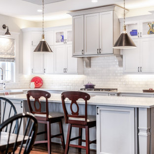 Modelo de cocina comedor en L, campestre, grande, con fregadero sobremueble, armarios estilo shaker, puertas de armario blancas, encimera de cuarzo compacto, salpicadero blanco, salpicadero de azulejos tipo metro, electrodomésticos de acero inoxidable, suelo de madera en tonos medios, una isla, suelo marrón, encimeras blancas y papel pintado