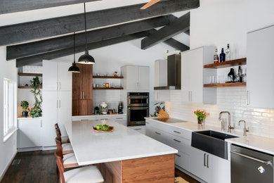 Enclosed kitchen - mid-sized mid-century modern galley laminate floor, brown floor and exposed beam enclosed kitchen idea in Denver with a farmhouse sink, flat-panel cabinets, white cabinets, solid surface countertops, white backsplash, subway tile backsplash, stainless steel appliances, an island and white countertops
