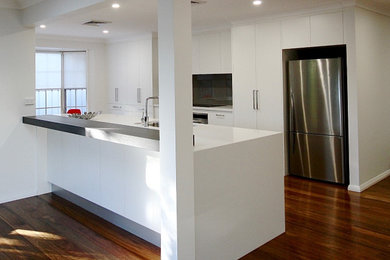 Modern single-wall open plan kitchen in Sydney with white cabinets, grey splashback, stainless steel appliances, dark hardwood floors, with island, brown floor and white benchtop.