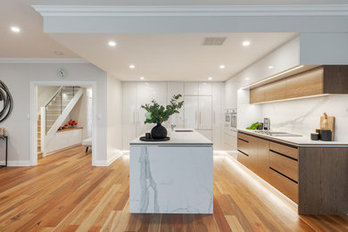 Mid-sized modern l-shaped open plan kitchen in Adelaide with an undermount sink, flat-panel cabinets, white cabinets, solid surface benchtops, white splashback, stone slab splashback, white appliances, light hardwood floors, with island, brown floor and white benchtop.
