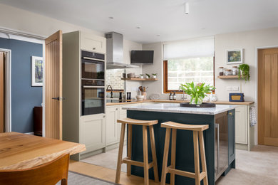 This is an example of a mid-sized country l-shaped eat-in kitchen in Gloucestershire with a single-bowl sink, beige cabinets, wood benchtops, black appliances, with island, beige floor and shaker cabinets.