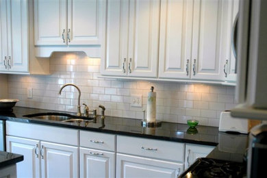 Example of a mid-sized classic l-shaped ceramic tile and black floor enclosed kitchen design in Other with a double-bowl sink, raised-panel cabinets, white cabinets, quartz countertops, white backsplash, cement tile backsplash, stainless steel appliances, an island and black countertops