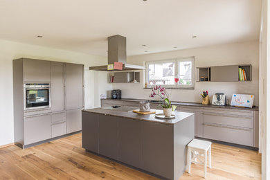 Große, Zweizeilige Moderne Wohnküche mit flächenbündigen Schrankfronten, grauen Schränken, Küchenrückwand in Weiß, Küchengeräten aus Edelstahl, braunem Holzboden, Kücheninsel, Unterbauwaschbecken, Granit-Arbeitsplatte, Kalk-Rückwand, braunem Boden und brauner Arbeitsplatte in Sonstige