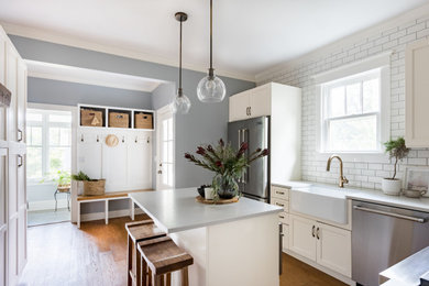 Example of a transitional light wood floor kitchen design in New York with a farmhouse sink, shaker cabinets, beige cabinets, white backsplash, ceramic backsplash, stainless steel appliances, an island and gray countertops