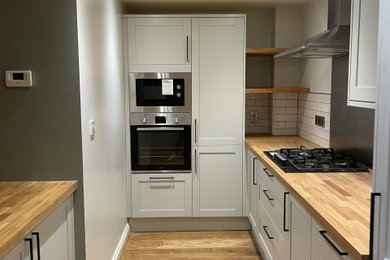 This is an example of a medium sized scandinavian grey and white kitchen/diner in West Midlands with a built-in sink, grey cabinets, wood worktops, white splashback, ceramic splashback, black appliances, light hardwood flooring, an island, brown floors and brown worktops.