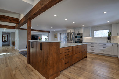 This is an example of a large transitional l-shaped kitchen in Other with a farmhouse sink, recessed-panel cabinets, white cabinets, quartz benchtops, white splashback, stainless steel appliances, light hardwood floors, with island, brown floor and white benchtop.