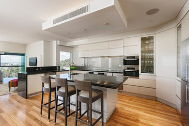 This is an example of a contemporary l-shaped eat-in kitchen in Perth with an undermount sink, flat-panel cabinets, beige cabinets, grey splashback, glass sheet splashback, stainless steel appliances, light hardwood floors and with island.