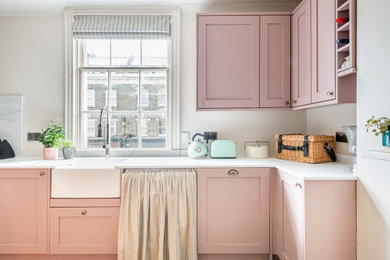 Design ideas for a medium sized contemporary u-shaped kitchen/diner in London with a belfast sink, shaker cabinets, pink cabinets, composite countertops, white splashback, vinyl flooring, an island, brown floors, white worktops and a chimney breast.