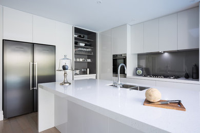 This is an example of a large contemporary l-shaped eat-in kitchen in Auckland with an undermount sink, flat-panel cabinets, grey cabinets, quartz benchtops, grey splashback, glass sheet splashback, stainless steel appliances, light hardwood floors, with island and beige floor.