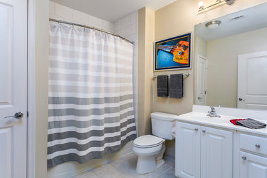 Photo of a mid-sized modern bathroom in Baltimore with raised-panel cabinets, white cabinets, an alcove tub, a shower/bathtub combo, a two-piece toilet, white tile, ceramic tile, ceramic floors, a drop-in sink, solid surface benchtops, beige floor and a shower curtain.