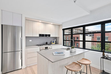 Inspiration for a small contemporary single-wall light wood floor and beige floor kitchen remodel in New York with an undermount sink, flat-panel cabinets, light wood cabinets, solid surface countertops, gray backsplash, stainless steel appliances, an island and gray countertops